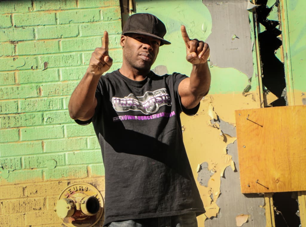 DaForce wearing a black cap and shirt in front of a green wall.